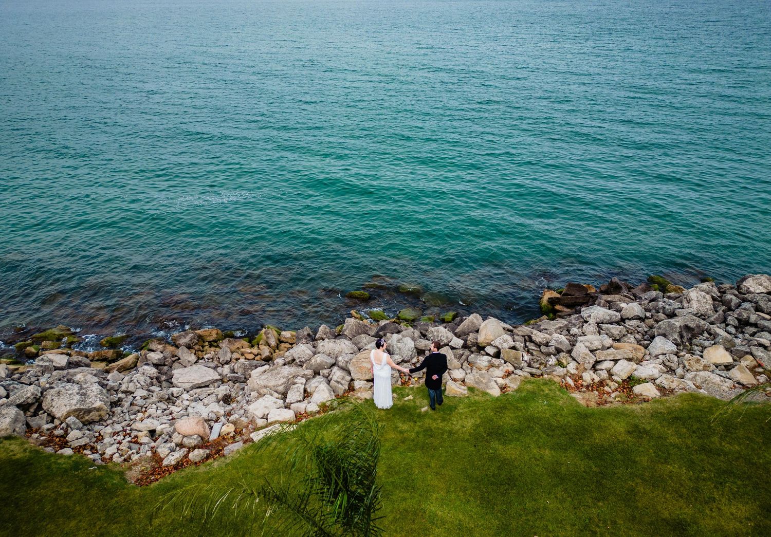 A couple stands on a grassy cliff overlooking turquoise waters and rocky shores.