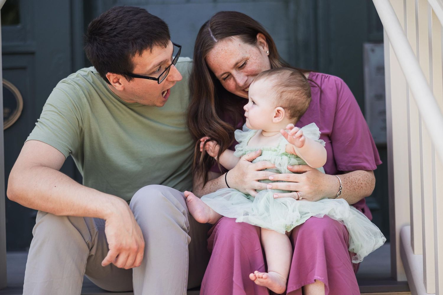A couple sitting on porch steps shares a tender moment with their baby wearing a white dress.