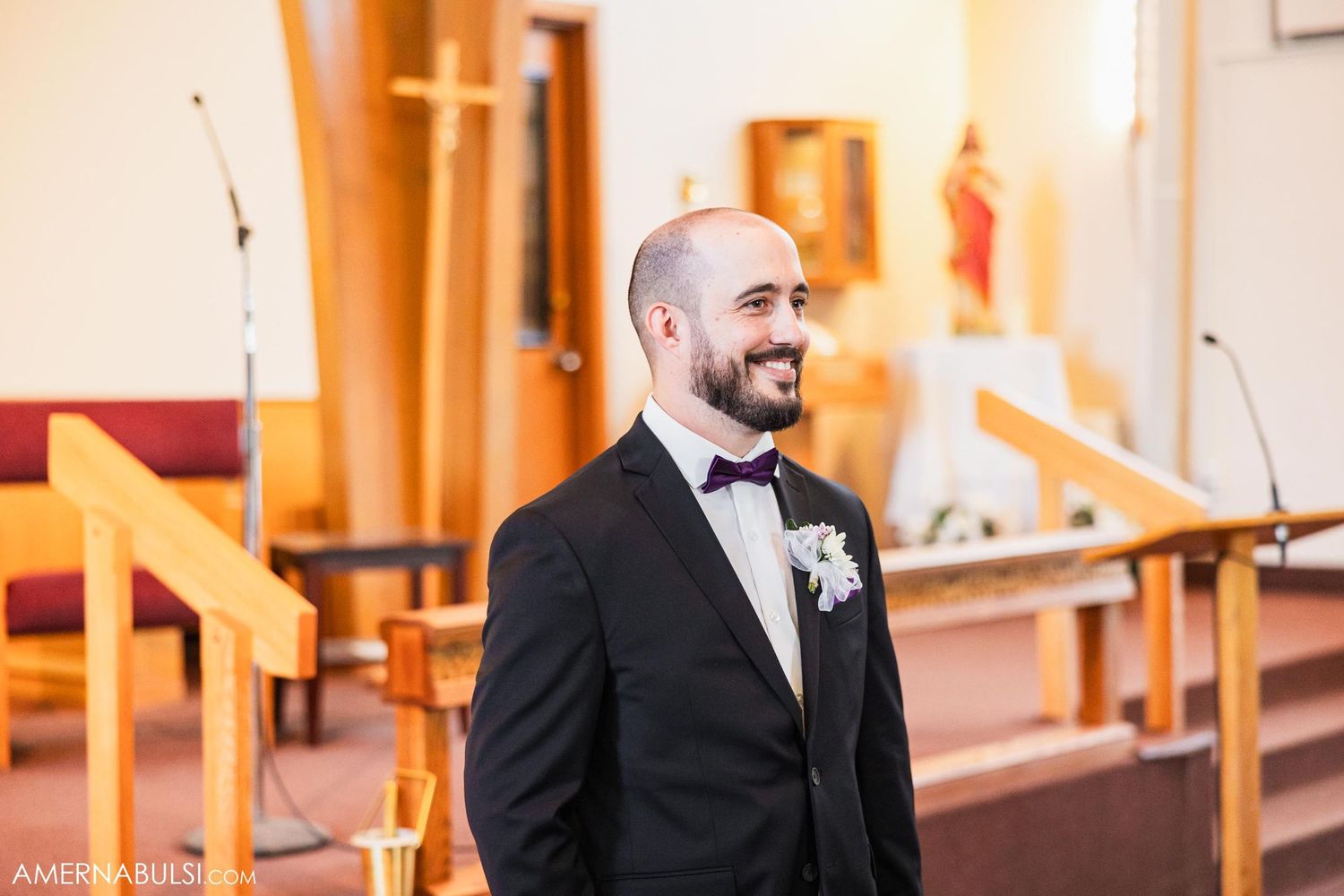 A groom in a black tuxedo and bowtie smiles warmly while standing in a church sanctuary before his wedding.