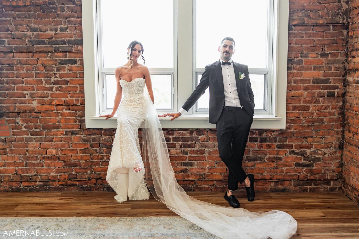 A couple poses by a bright window in an industrial-style wedding venue with exposed brick walls.