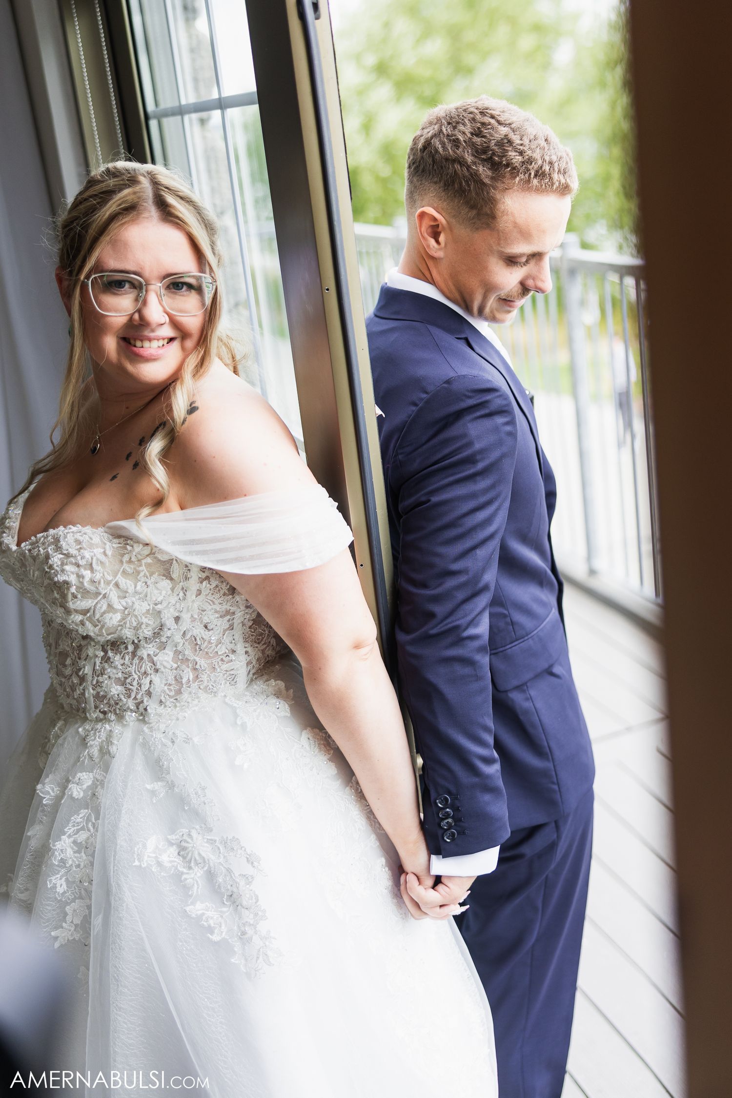 A bride in a lace wedding gown poses near windows.