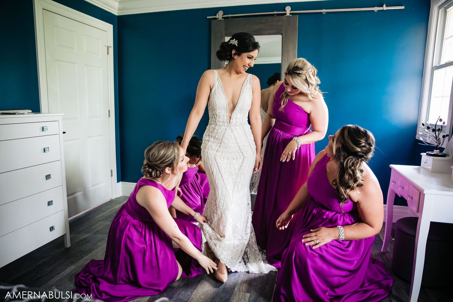 Bridesmaids in magenta dresses help adjust a wedding gown in a blue room.