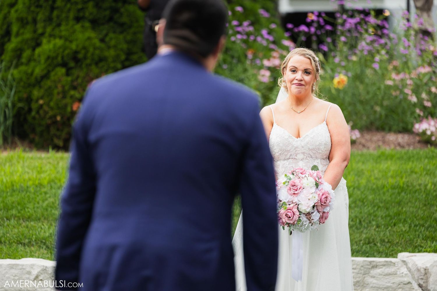 A bride in white holds a pink and white bouquet while looking at someone in a navy suit during an outdoor ceremony.