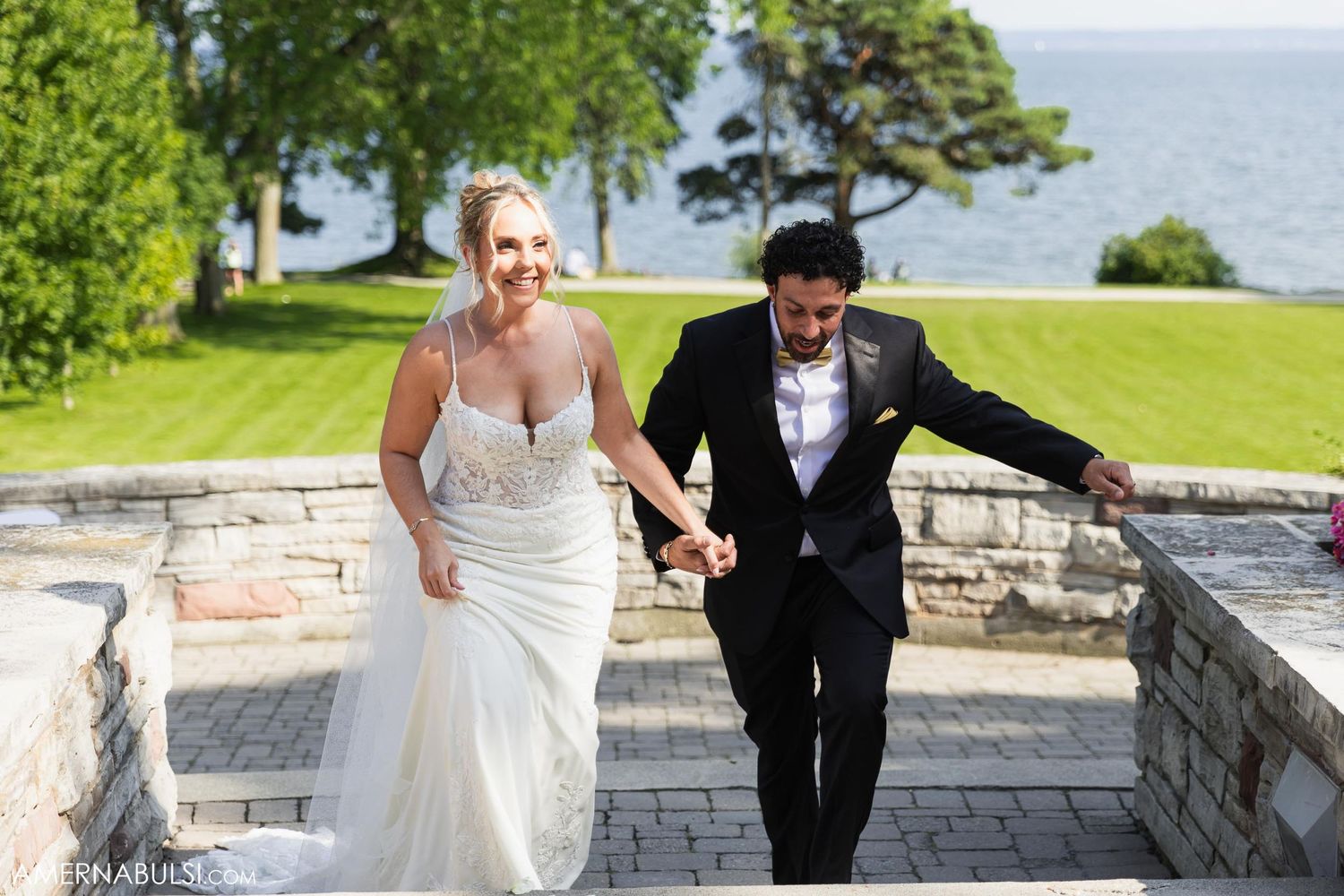 A newly married couple walking together and smiling on stone steps with waterfront views.