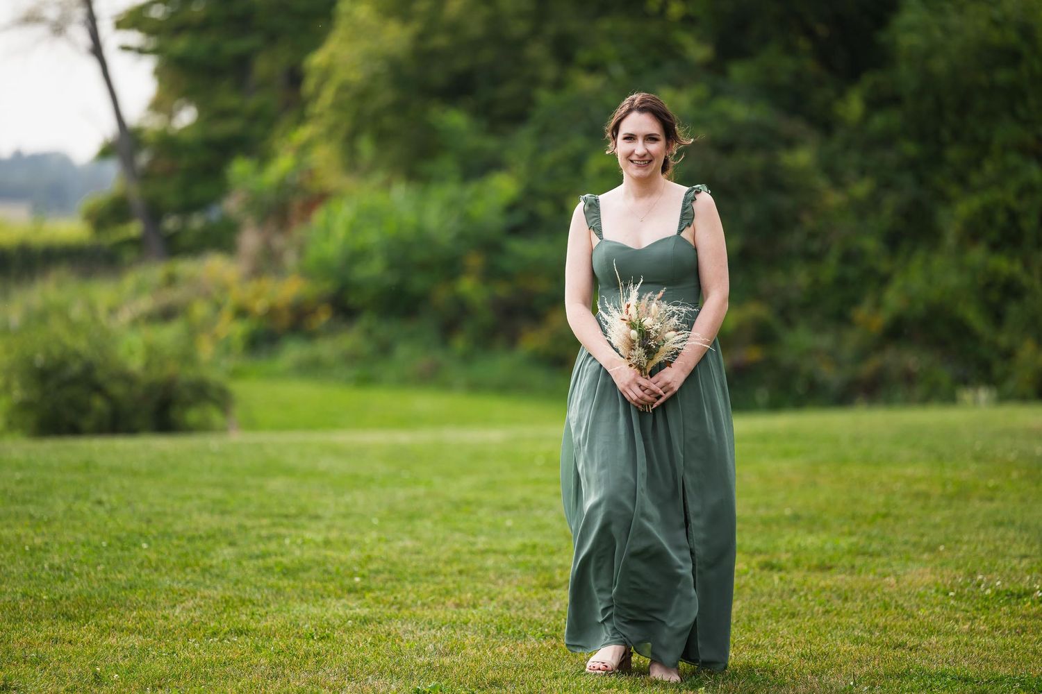 Bridesmaid in long green dress holds bouquet while standing on grass.