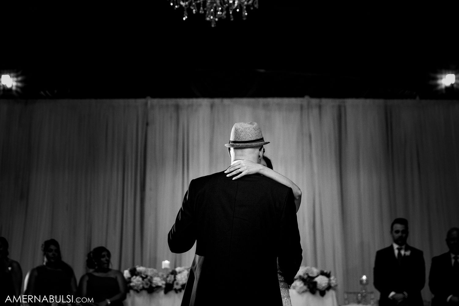 A black and white photo of a performer in a hat and suit facing a curtained stage at a formal event.