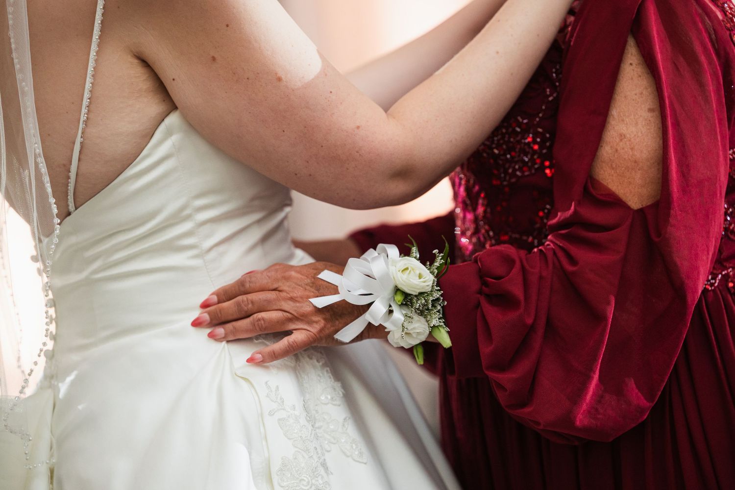 Bride in white lace gown embraced by woman in red velvet dress holding white flower.