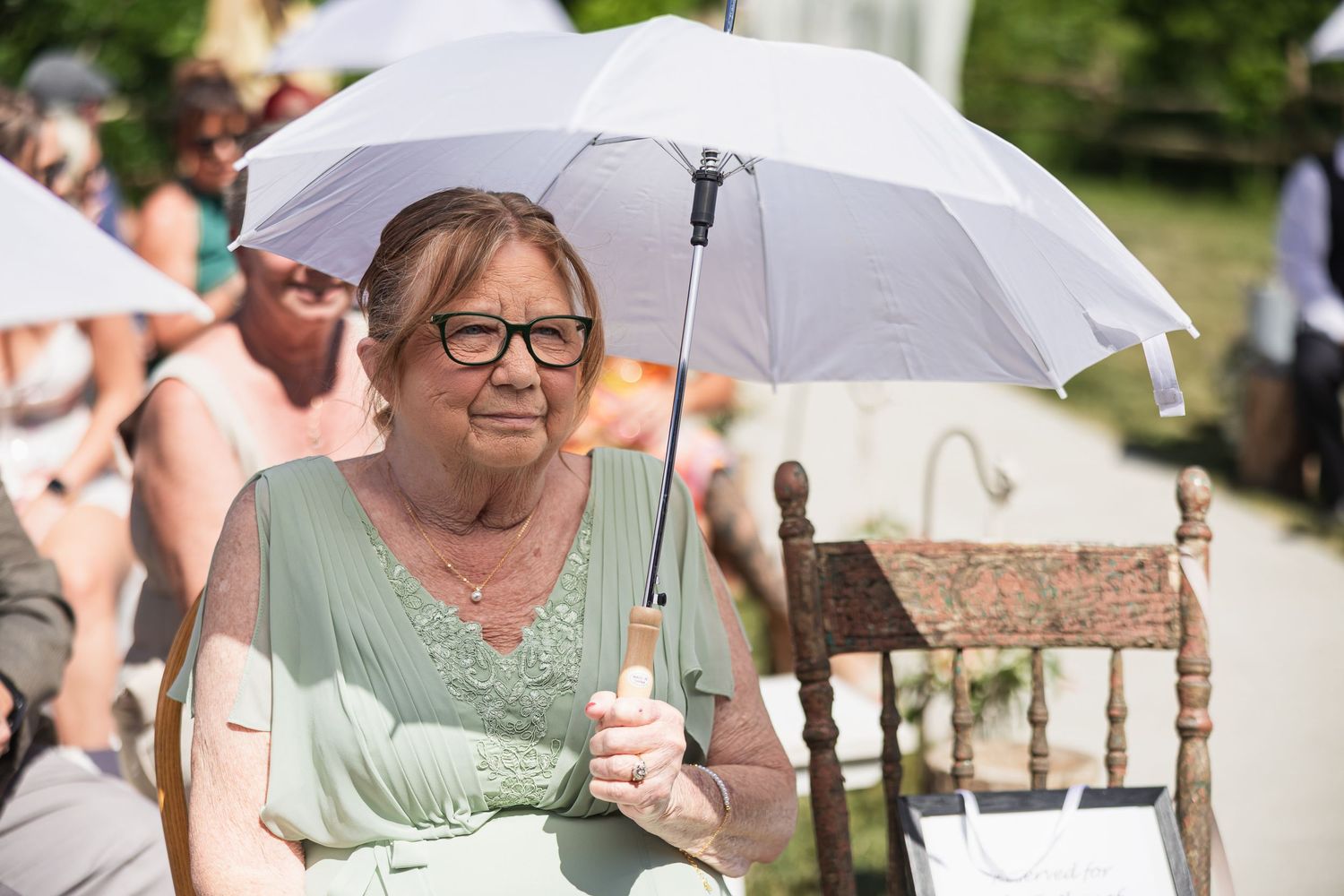 Elderly woman wearing glasses holding a white parasol umbrella outdoors at a sunny wedding ceremony.