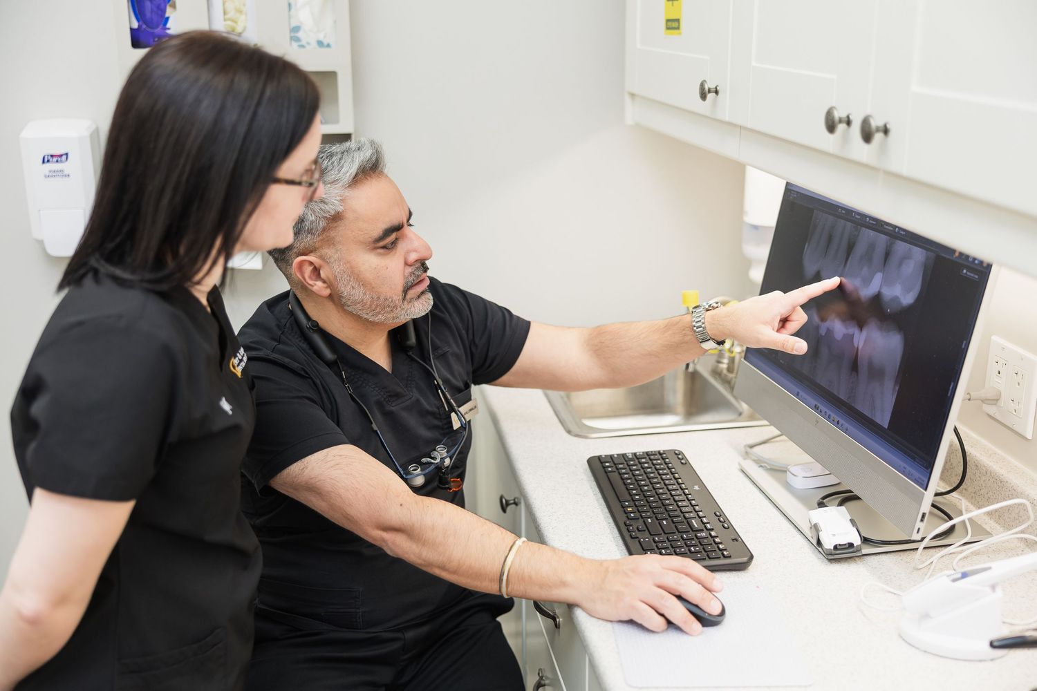 Two dental professionals in black scrubs reviewing dental X-rays on a computer monitor in a clinical setting.