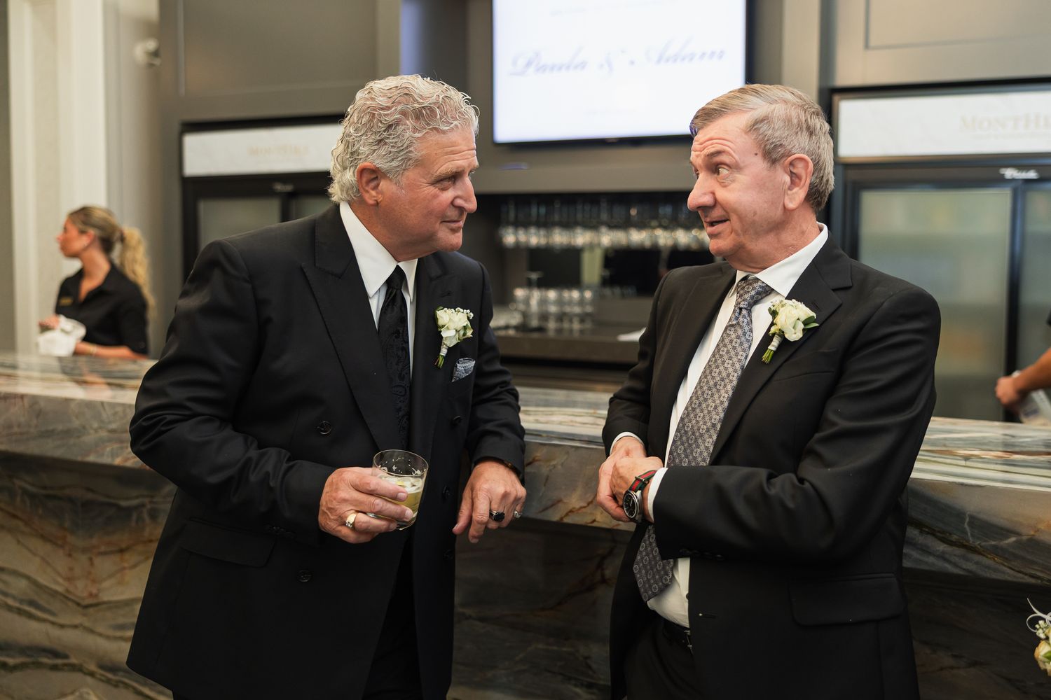 Two men in black suits chatting and smiling at a wedding reception near a marble bar.