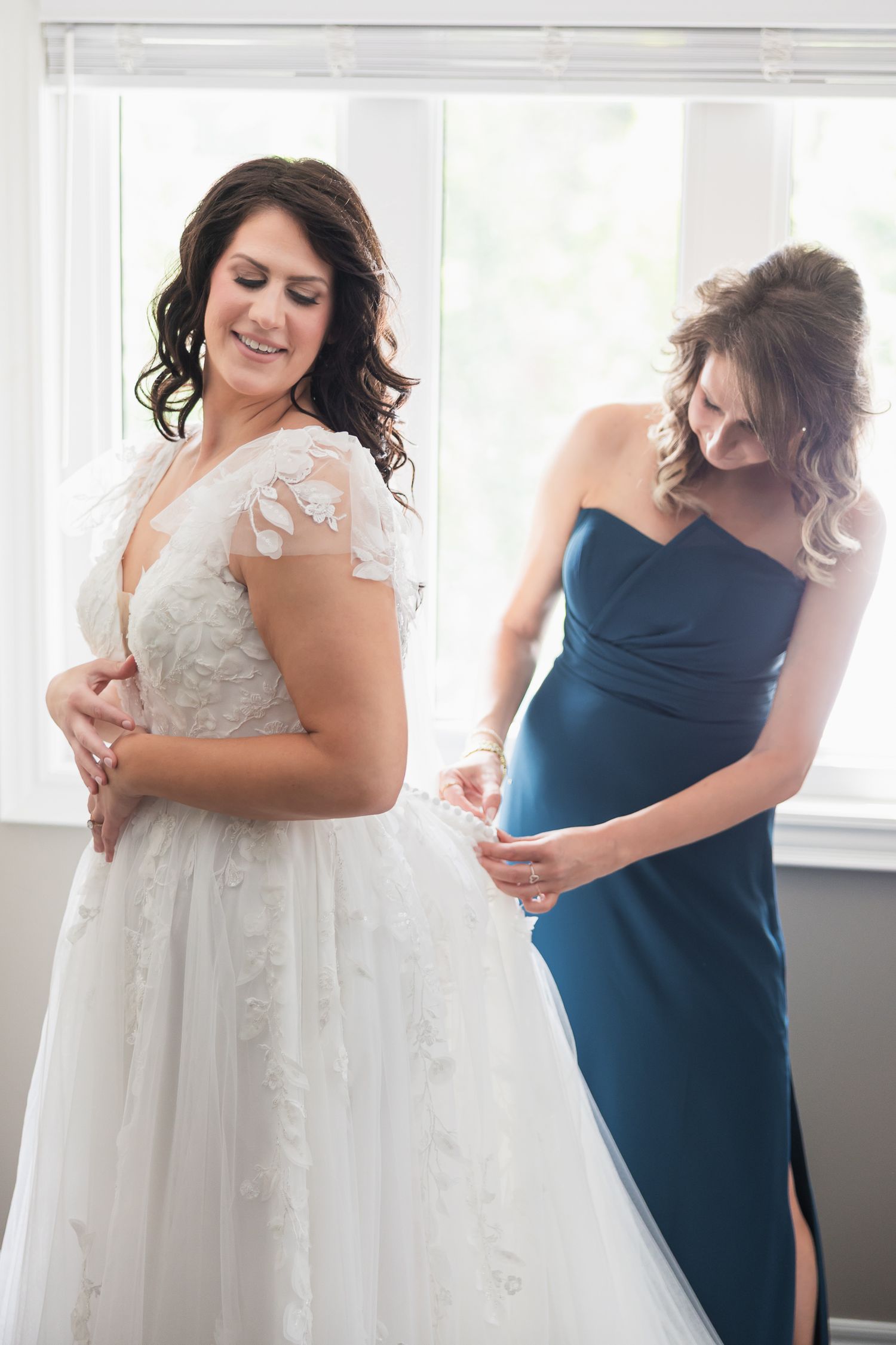 Bride in lace wedding gown being helped by bridesmaid in blue dress near bright window.