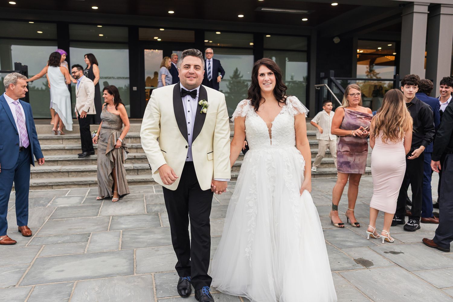 Bride and groom walking hand in hand outside a wedding venue, surrounded by elegantly dressed guests on steps.
