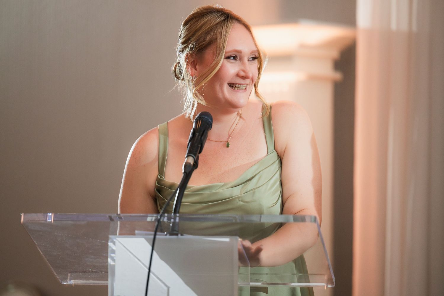 Woman in green satin dress smiling at clear podium with microphone during formal event.