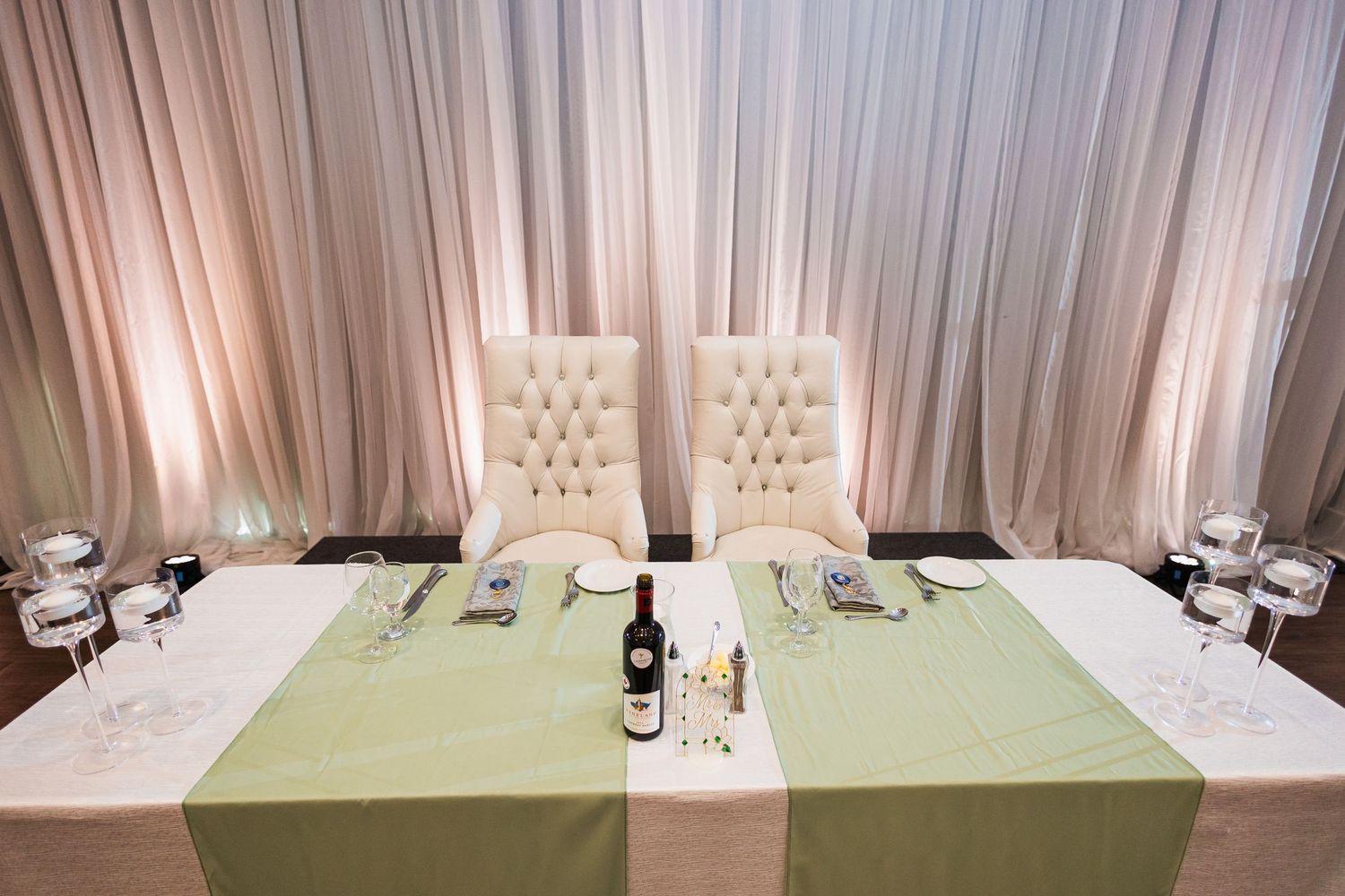 Elegant sweetheart table with tufted ivory chairs, green runner, wine bottle, and crystal glassware at wedding reception.