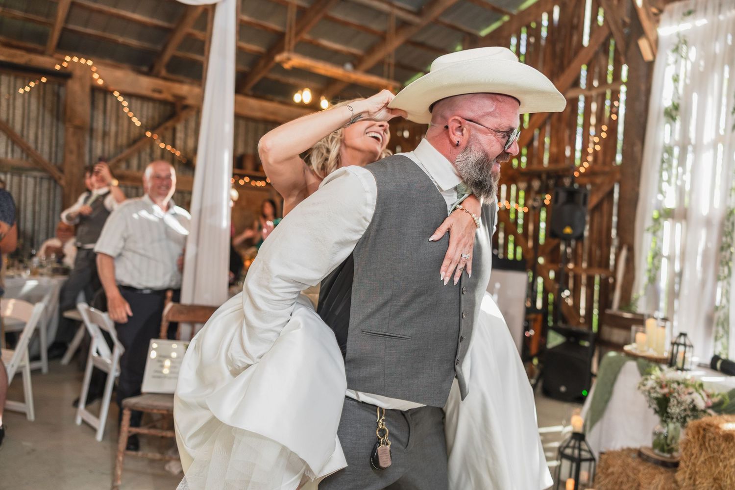 Bride and groom share first dance in rustic barn wedding venue, bride touching groom's white cowboy hat.