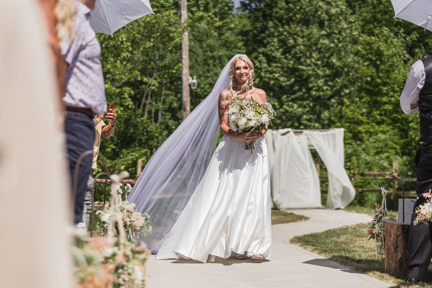 Smiling bride walks down outdoor aisle in white gown holding bouquet on sunny wedding day.