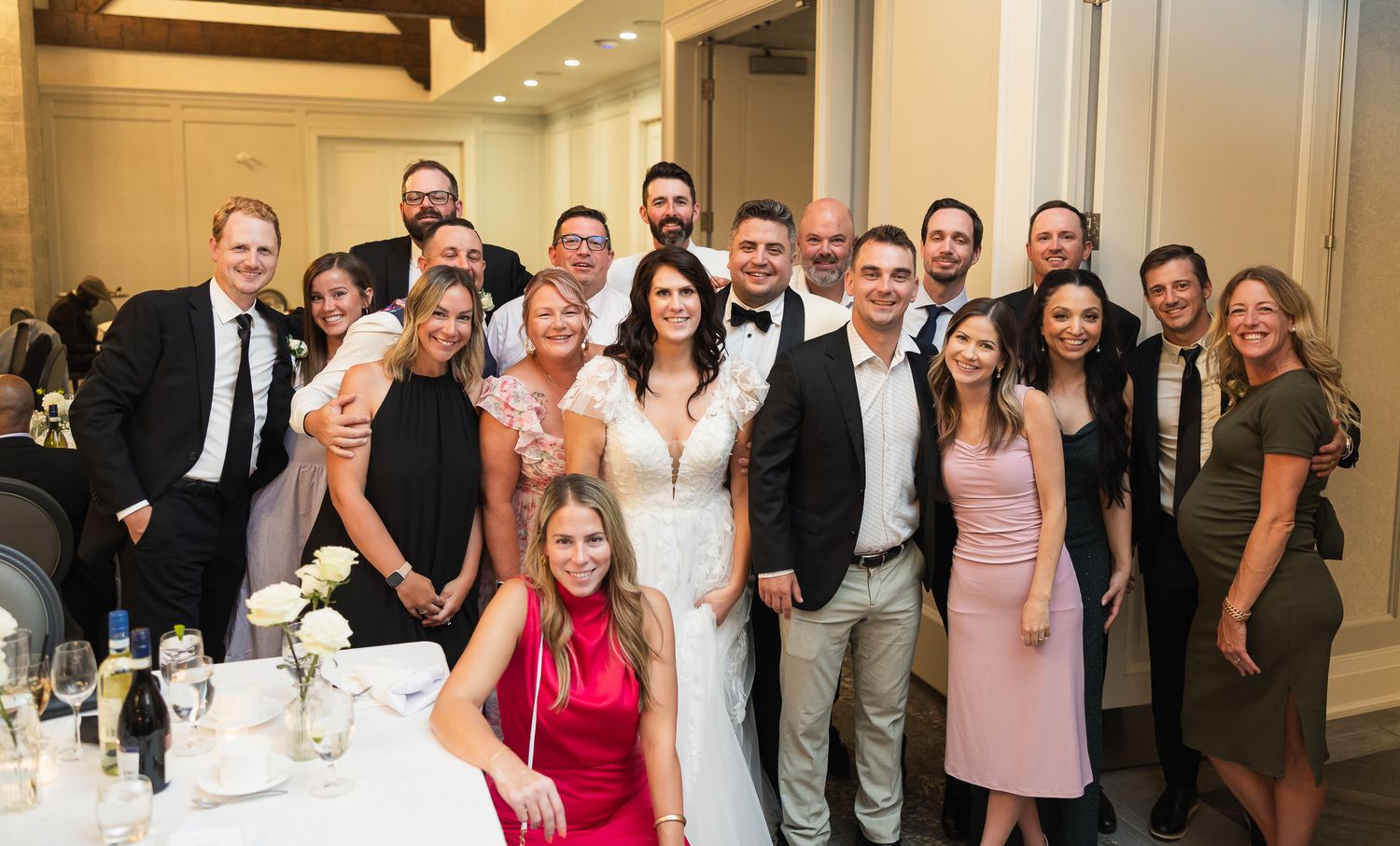 Wedding couple poses with guests in elegant ballroom reception venue for group celebration photo.