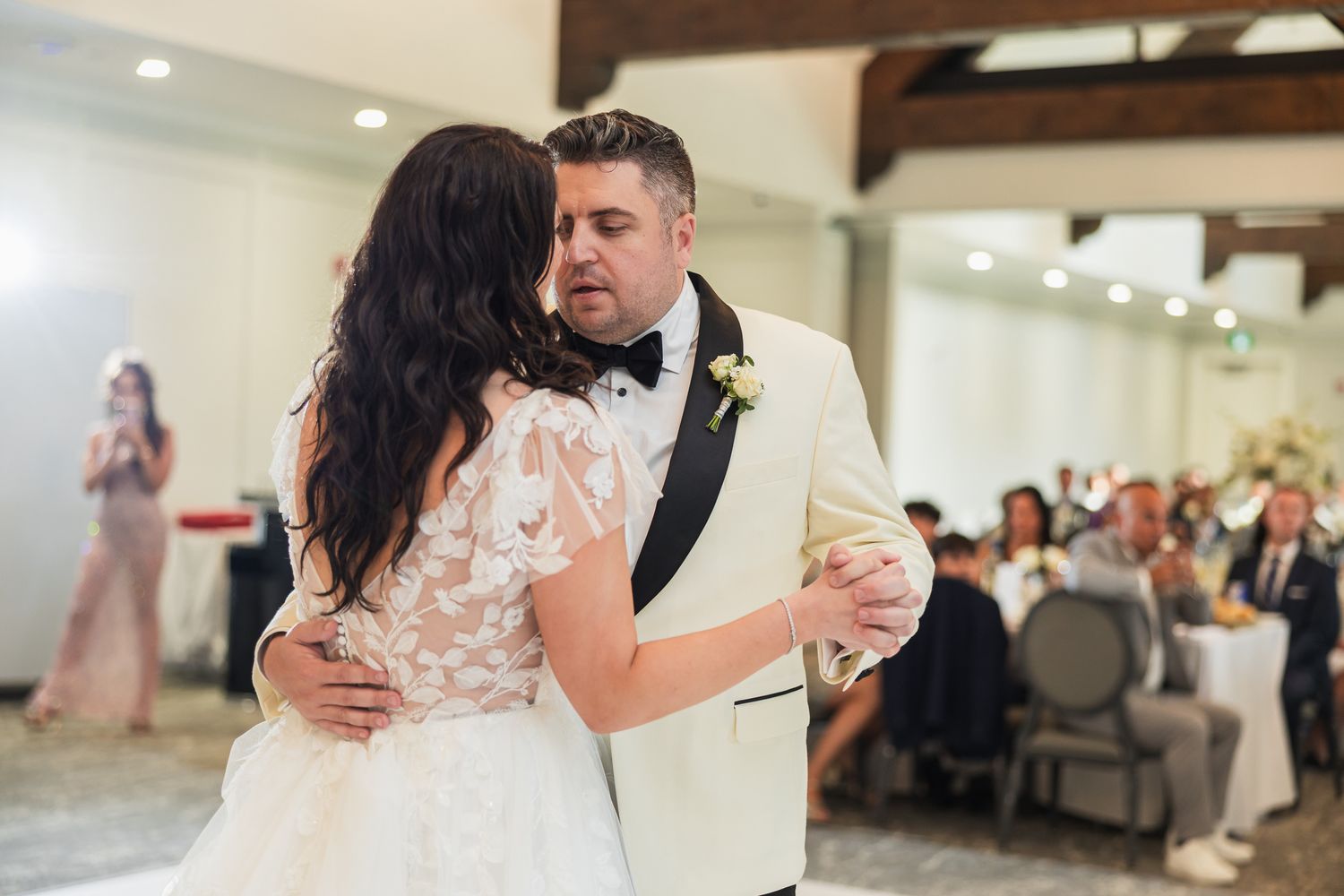 Bride and groom share their first dance at elegant wedding reception, groom in white tuxedo, bride in lace gown.
