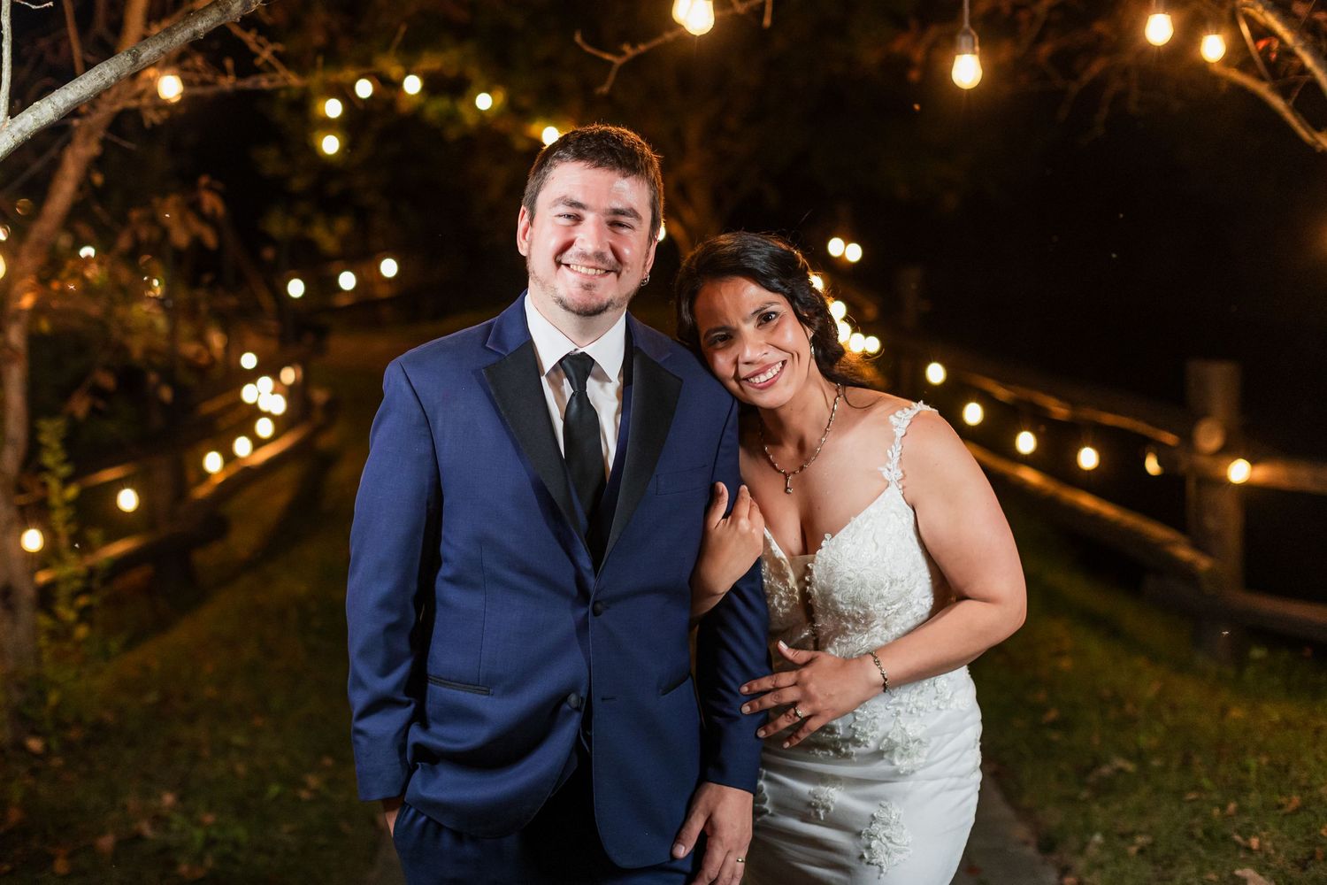 Smiling bride and groom pose together outdoors at night with warm string lights glowing behind them.
