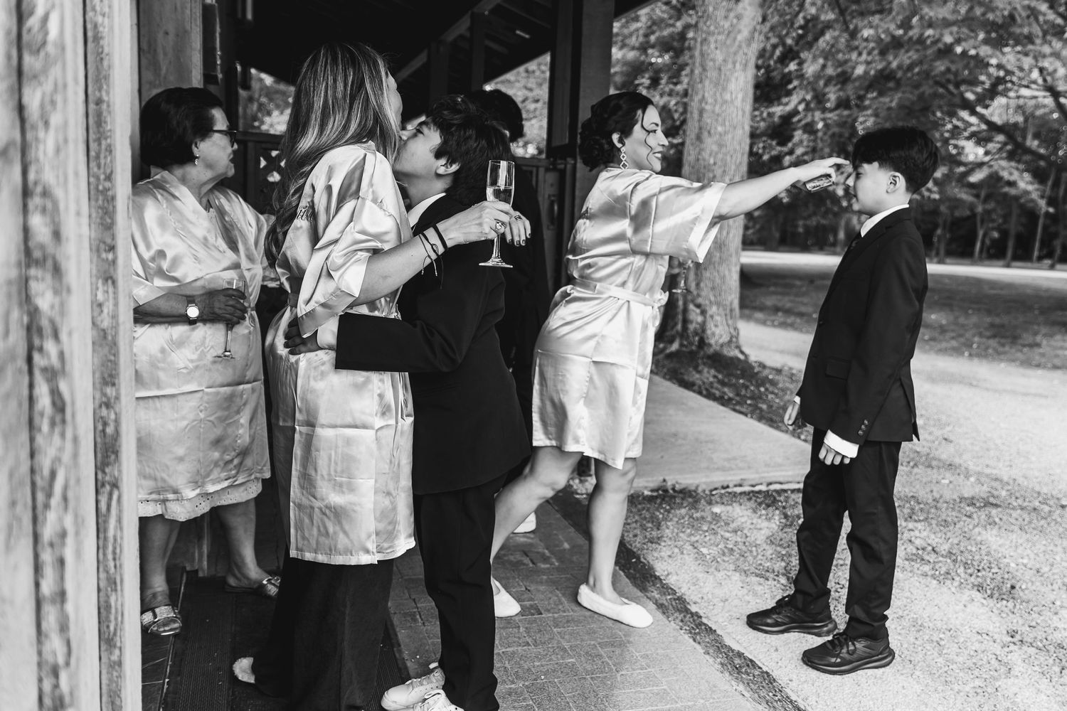 Bridesmaids in satin robes playfully interacting with young boy in suit in black and white photo.