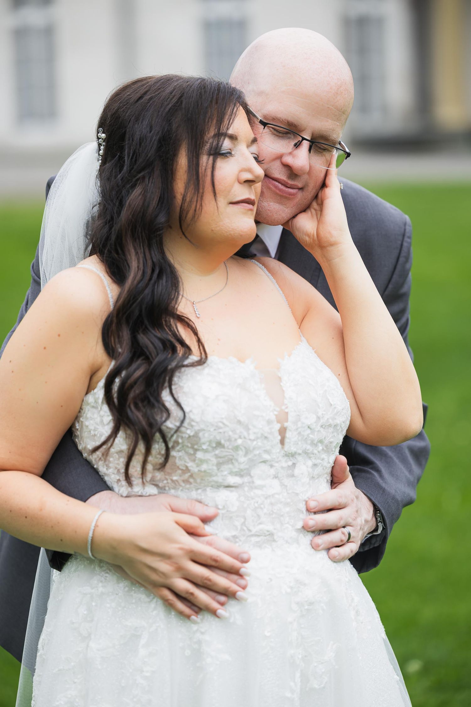 A tender wedding moment between newlyweds embracing on a grassy lawn in front of an elegant building.