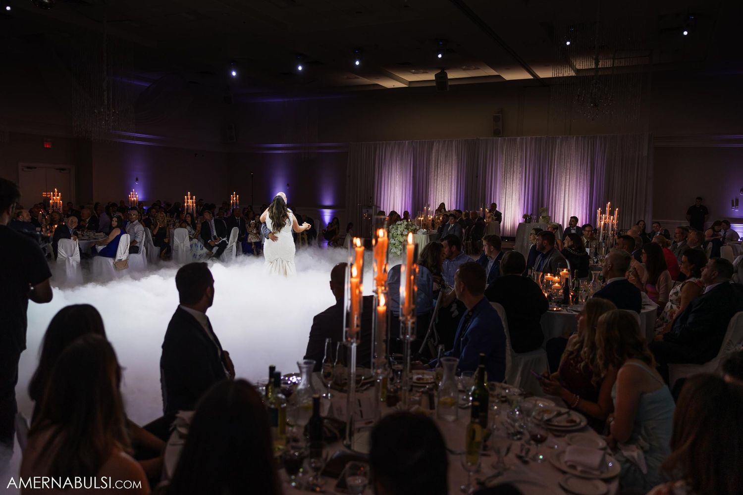 Wedding reception venue with low lighting, candles and purple uplighting while guests dance on foggy dancefloor.