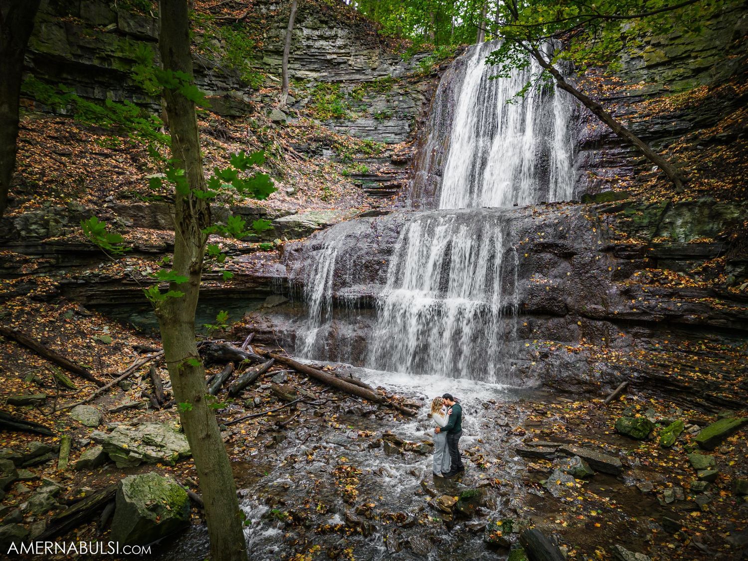 An aerial view of a waterfall in the woods with tiny figures standing at its base.