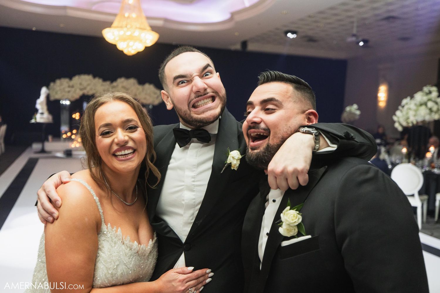 Wedding couple poses for candid group photo in formal attire under elegant chandelier lighting.
