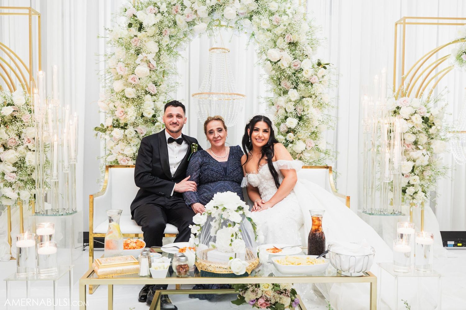Wedding party poses together on elegant white and gold furniture with floral arch backdrop.