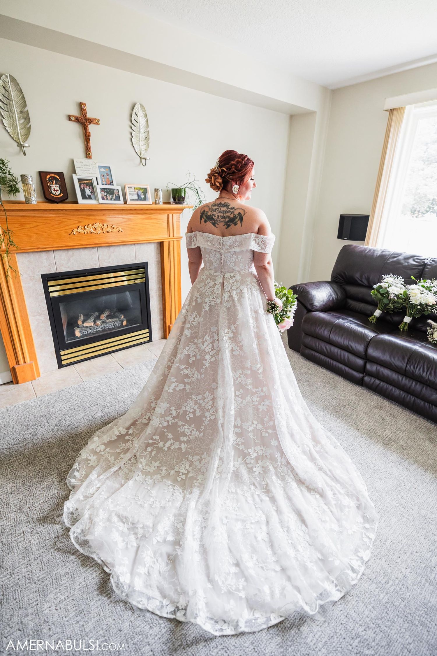 Lace wedding dress with long train shown from behind in living room with fireplace.