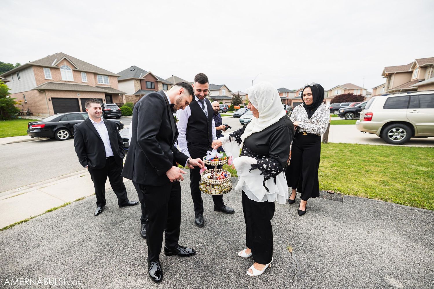 Wedding party members in formal attire gather and interact on driveway outside suburban home.
