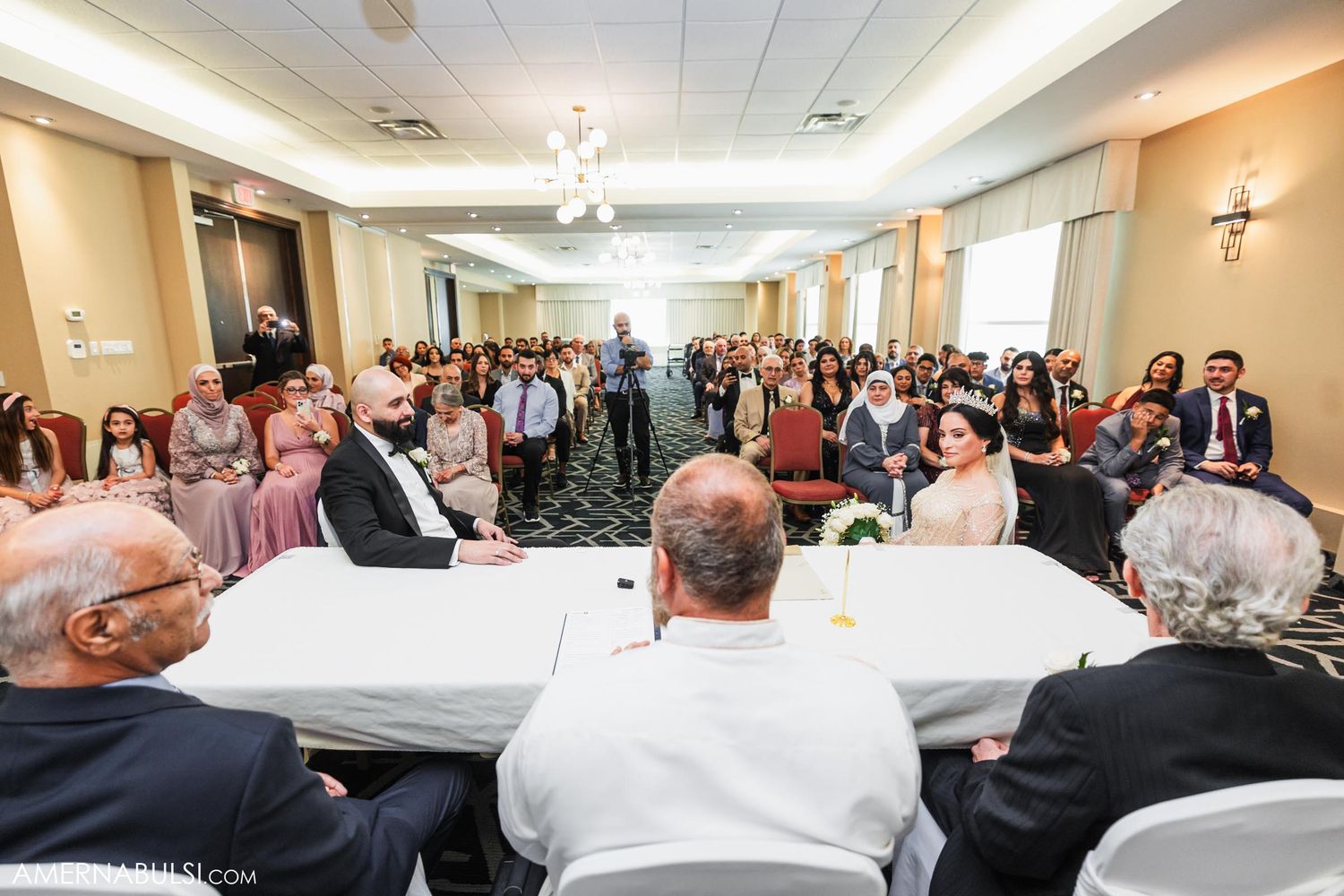 Wedding guests gather in a banquet hall for a ceremony with rows of chairs and elegant lighting.