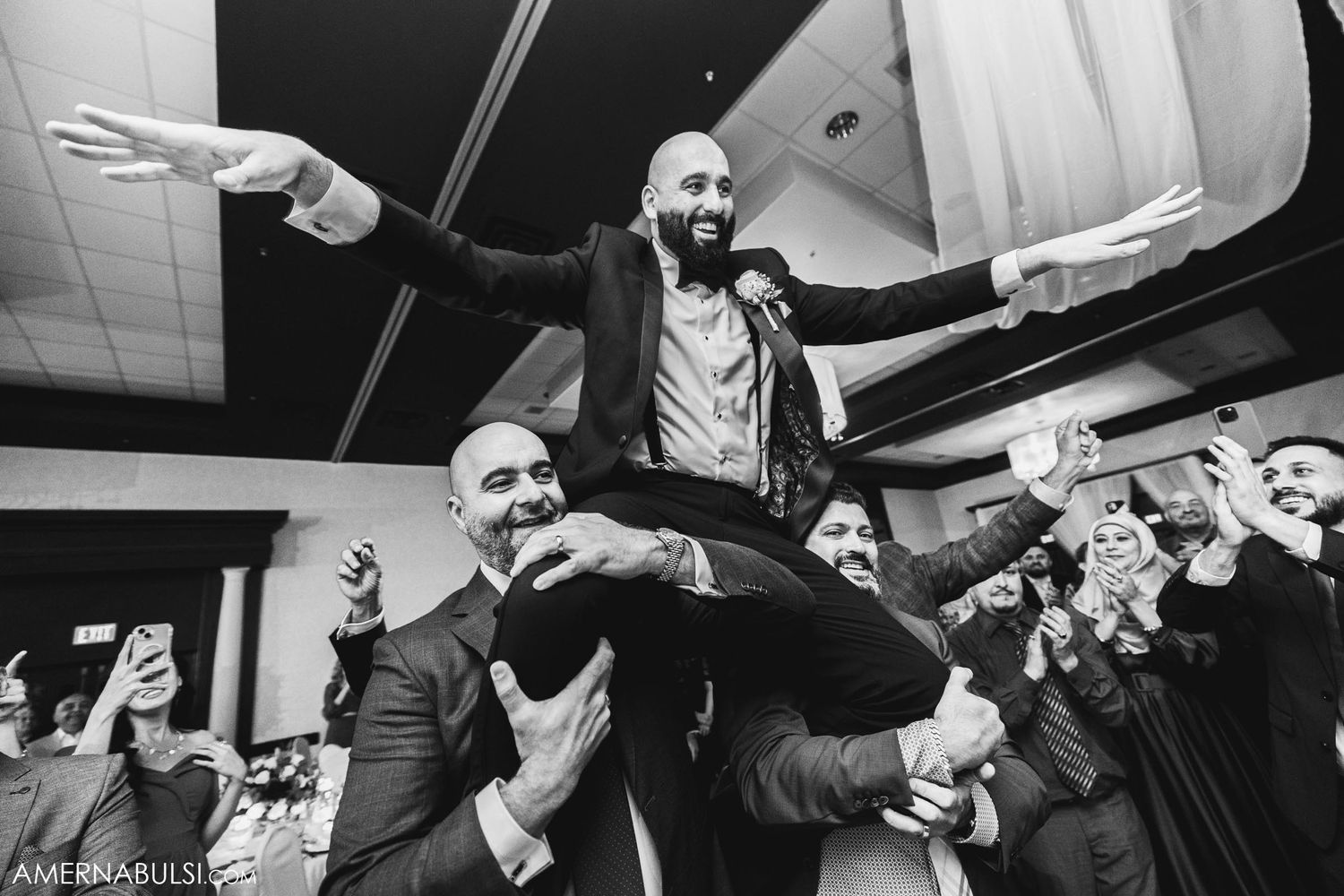 A groom being lifted by guests during lively wedding reception celebrations.