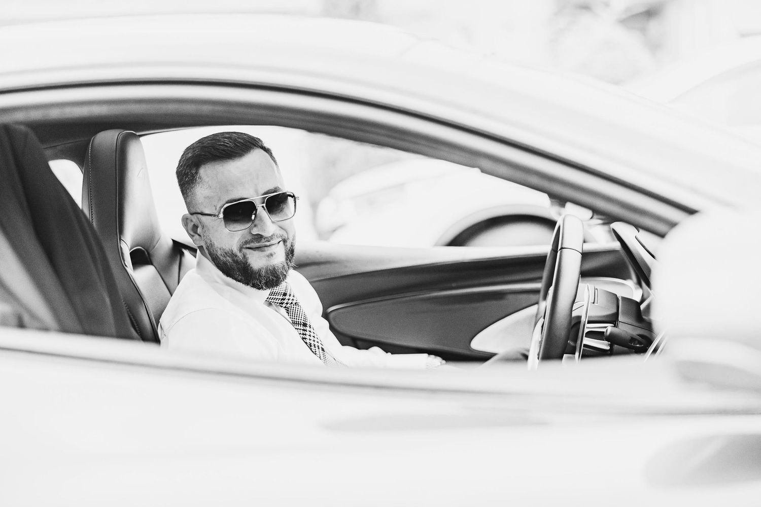 Black and white portrait through car window showing person wearing sunglasses behind steering wheel.