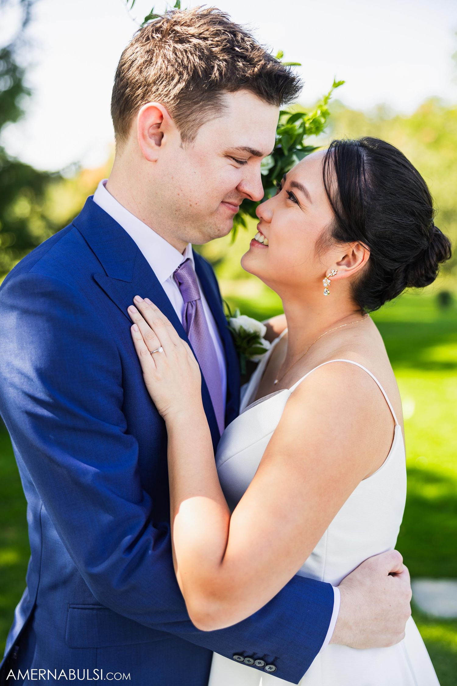 Bride and groom share an intimate moment in a romantic outdoor wedding photoshoot wearing blue suit and white dress.