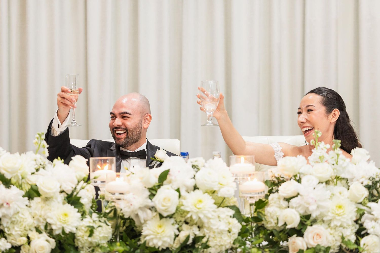 Wedding couple celebrating at head table decorated with white floral arrangements.