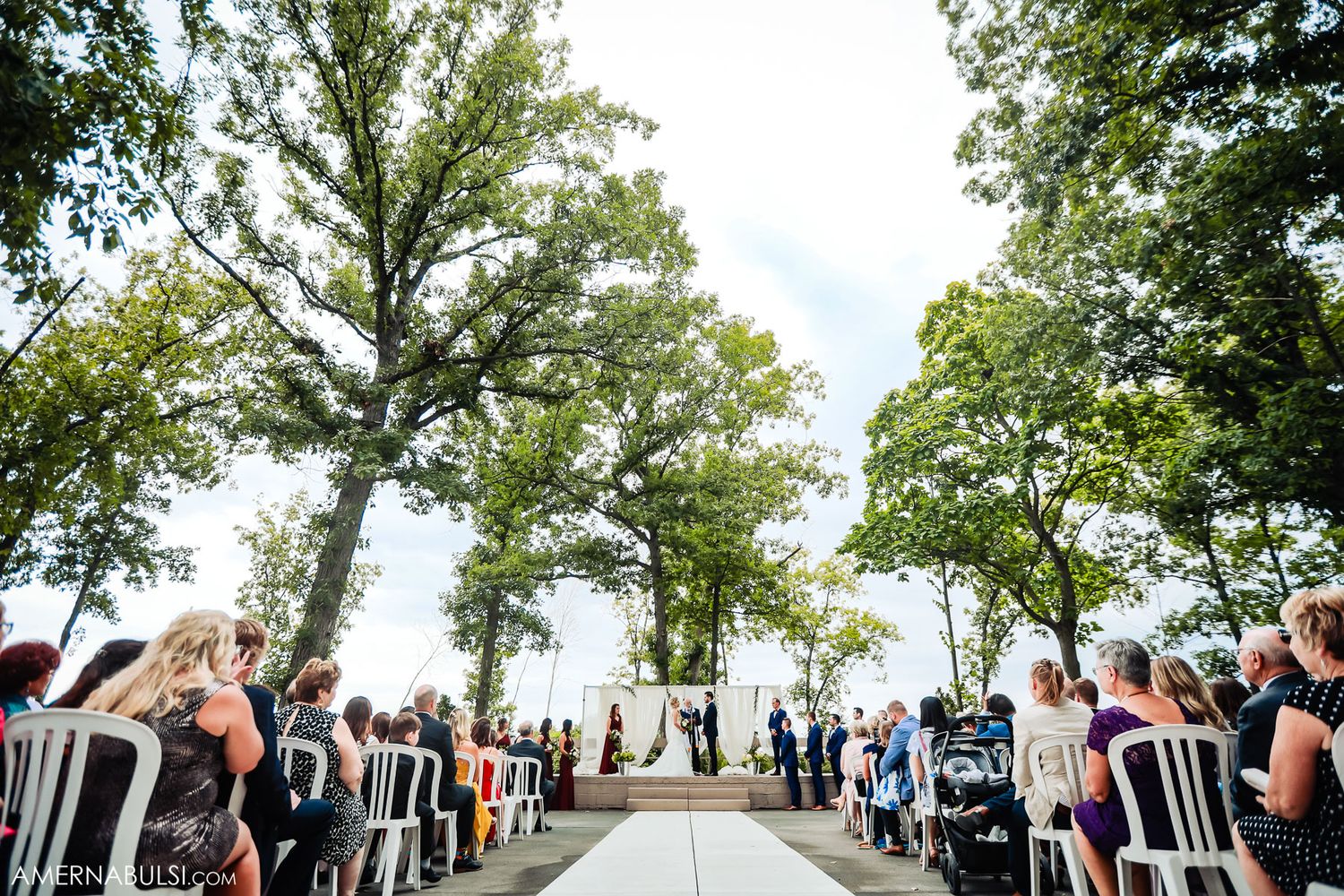 Outdoor wedding ceremony setup with white chairs along a tree-lined aisle overlooking a scenic waterfront view.