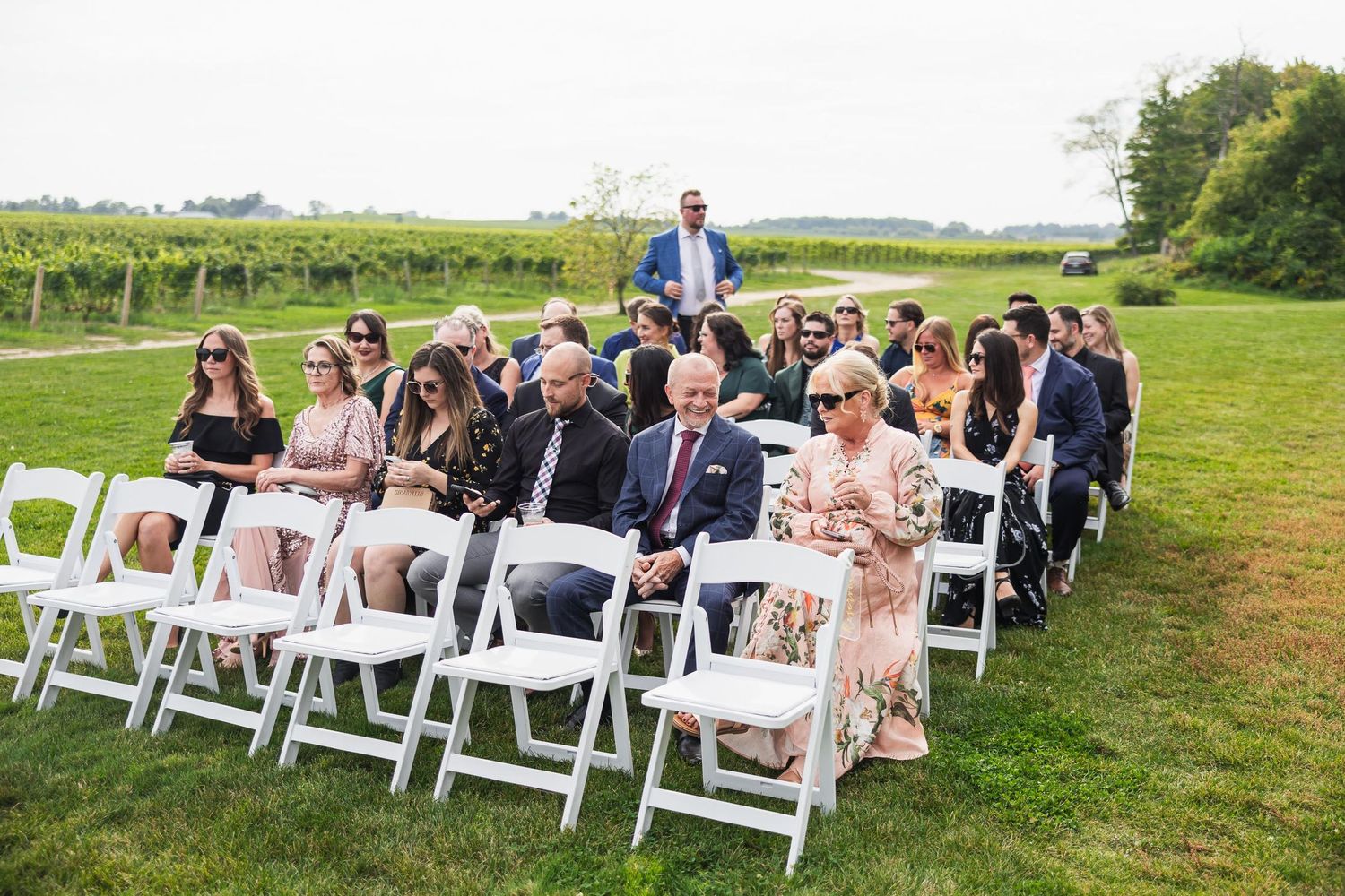 Wedding guests seated in white chairs on lawn with vineyard in background.