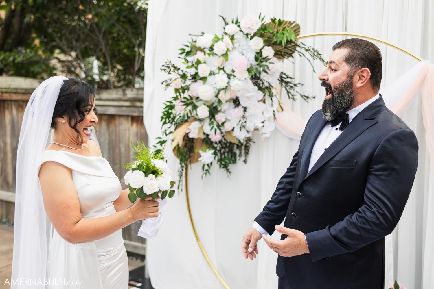 Bride and groom share candid moment in front of white floral wedding arch.