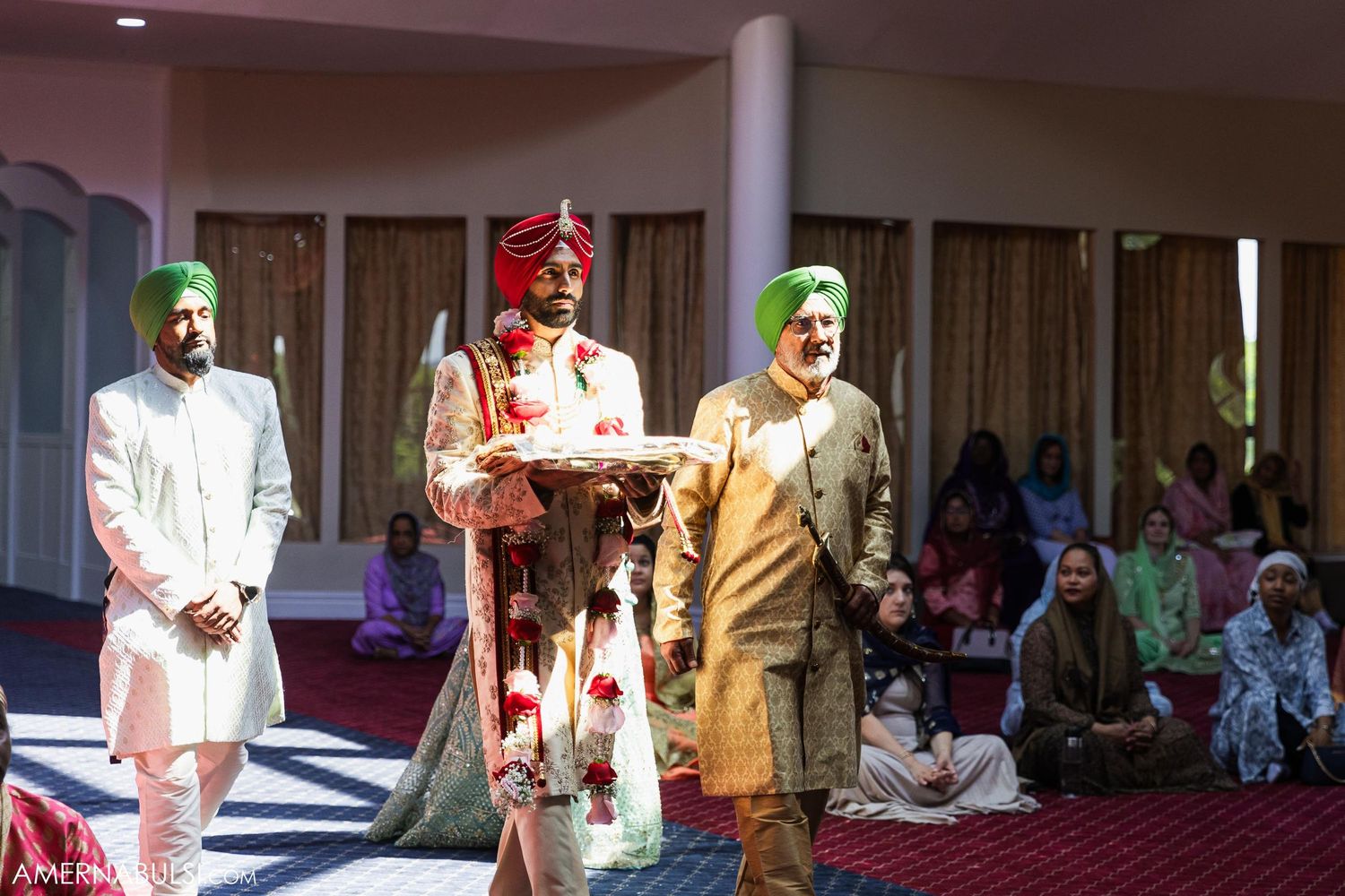 Sikh wedding ceremony participants in traditional attire standing in an indoor venue with gathered guests.