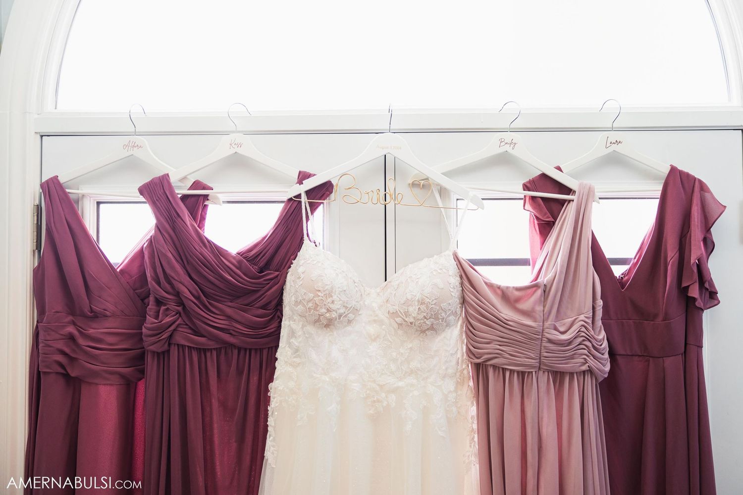 Wedding dresses and burgundy bridesmaid dresses hanging on white hangers against a white wall.
