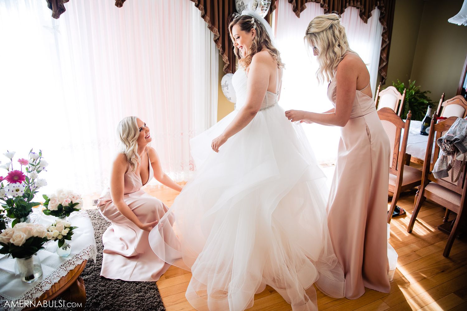 Bridesmaids in blush pink dresses helping with wedding preparations in a sunlit room.