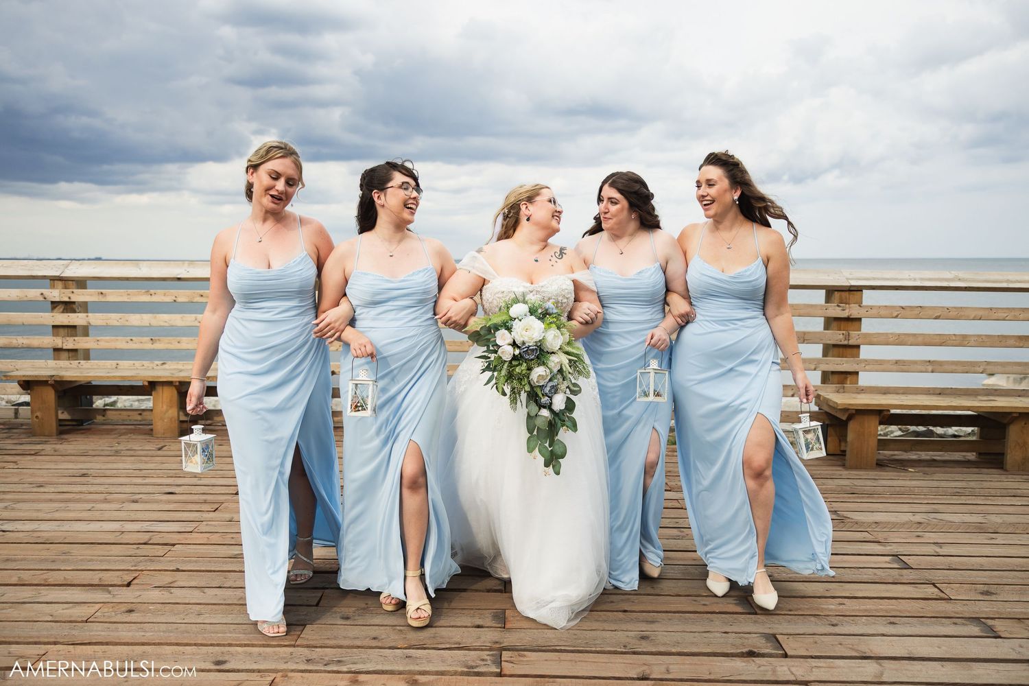 Bridal party in light blue dresses posing together on wooden boardwalk by the ocean.