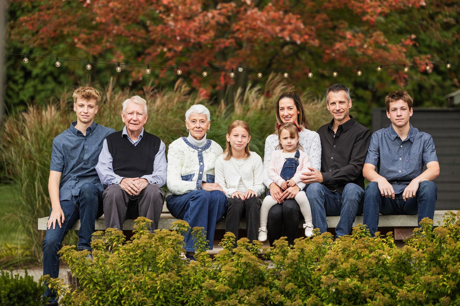 A group sits together outdoors on a bench surrounded by autumn foliage and greenery in a park setting.