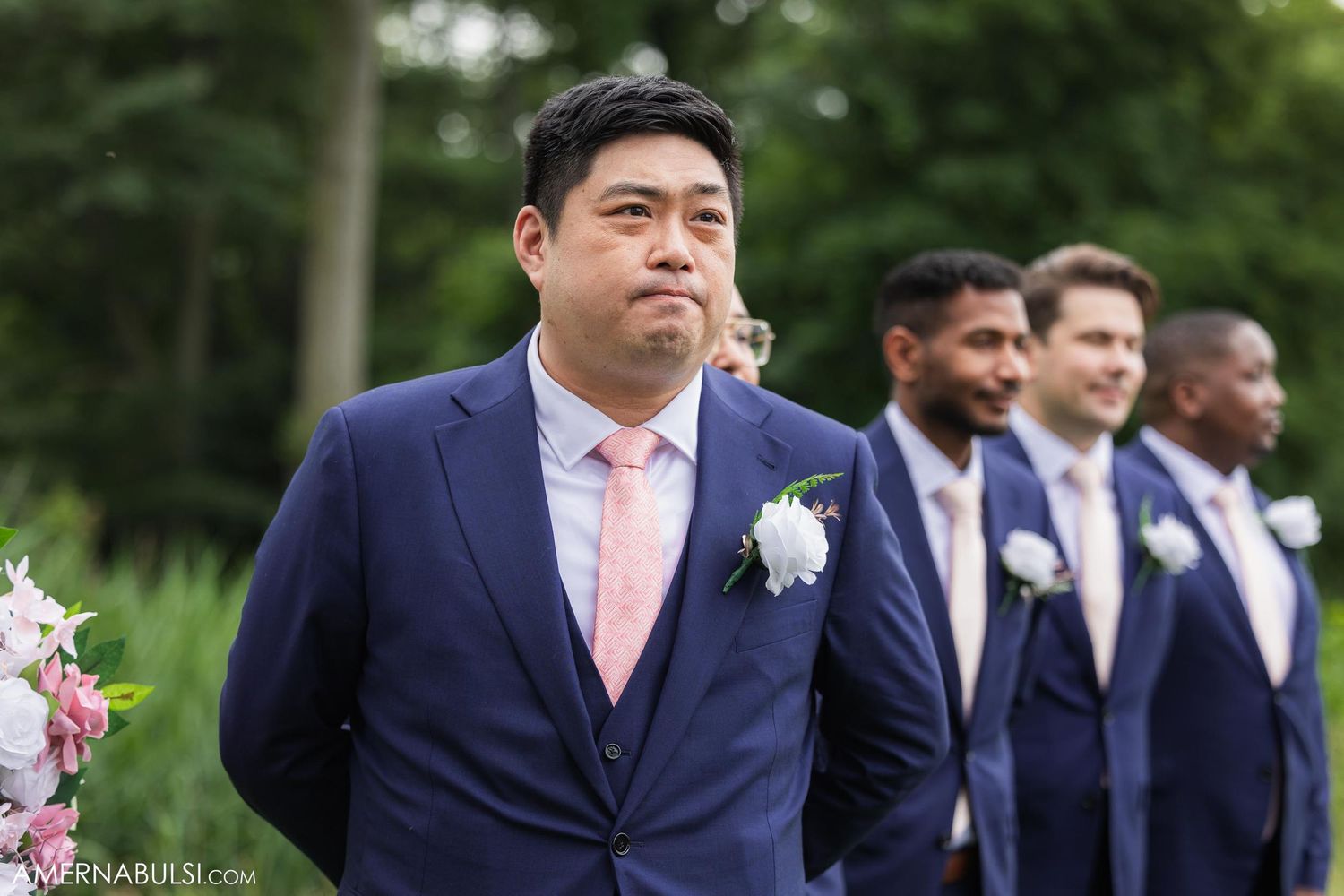 Groomsmen in navy suits with pink accents stand in line during outdoor wedding ceremony.