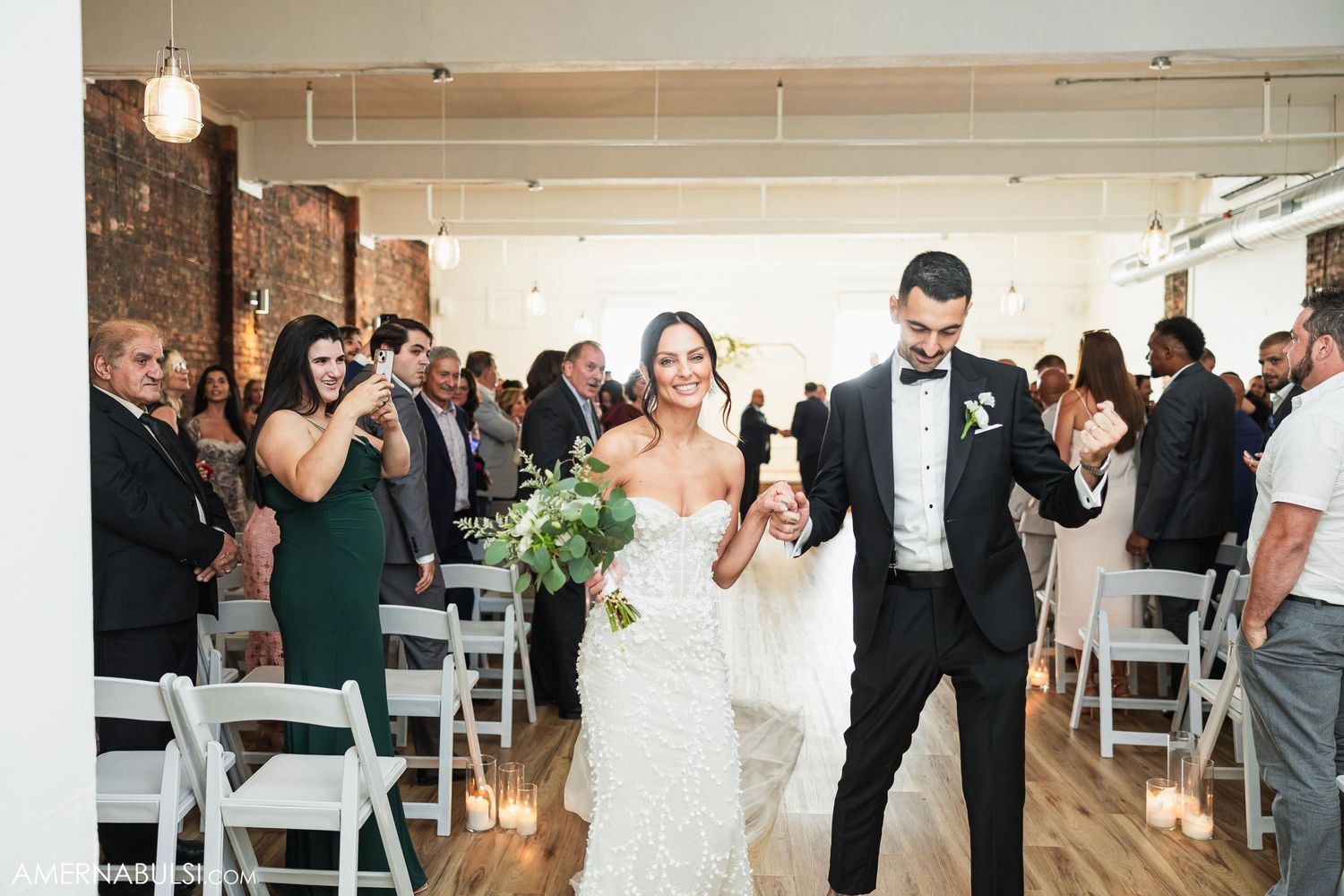 Wedding ceremony recessional in a bright venue with white chairs and exposed brick walls.