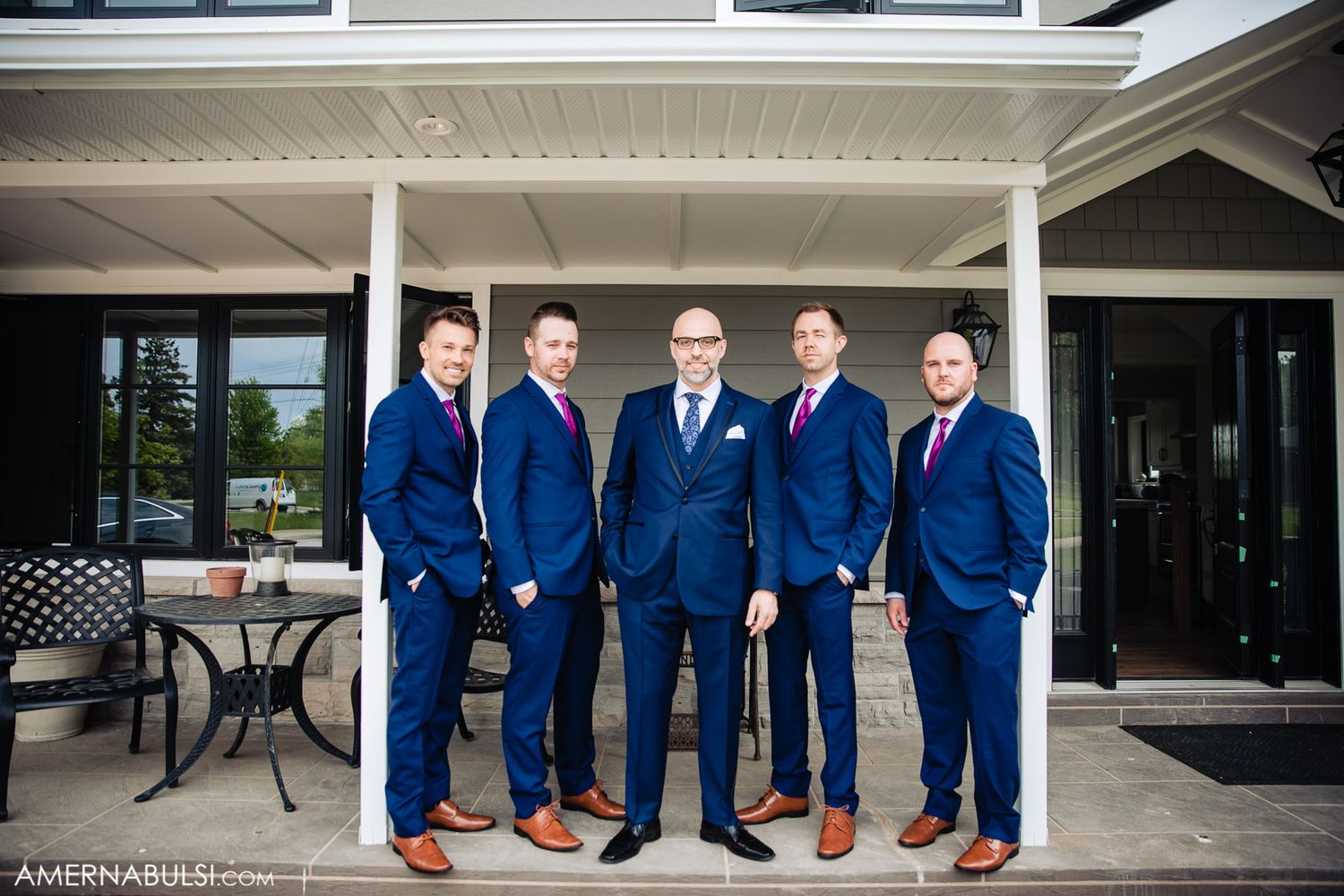 Groomsmen in matching navy blue suits and brown shoes pose together on a porch for wedding photos.