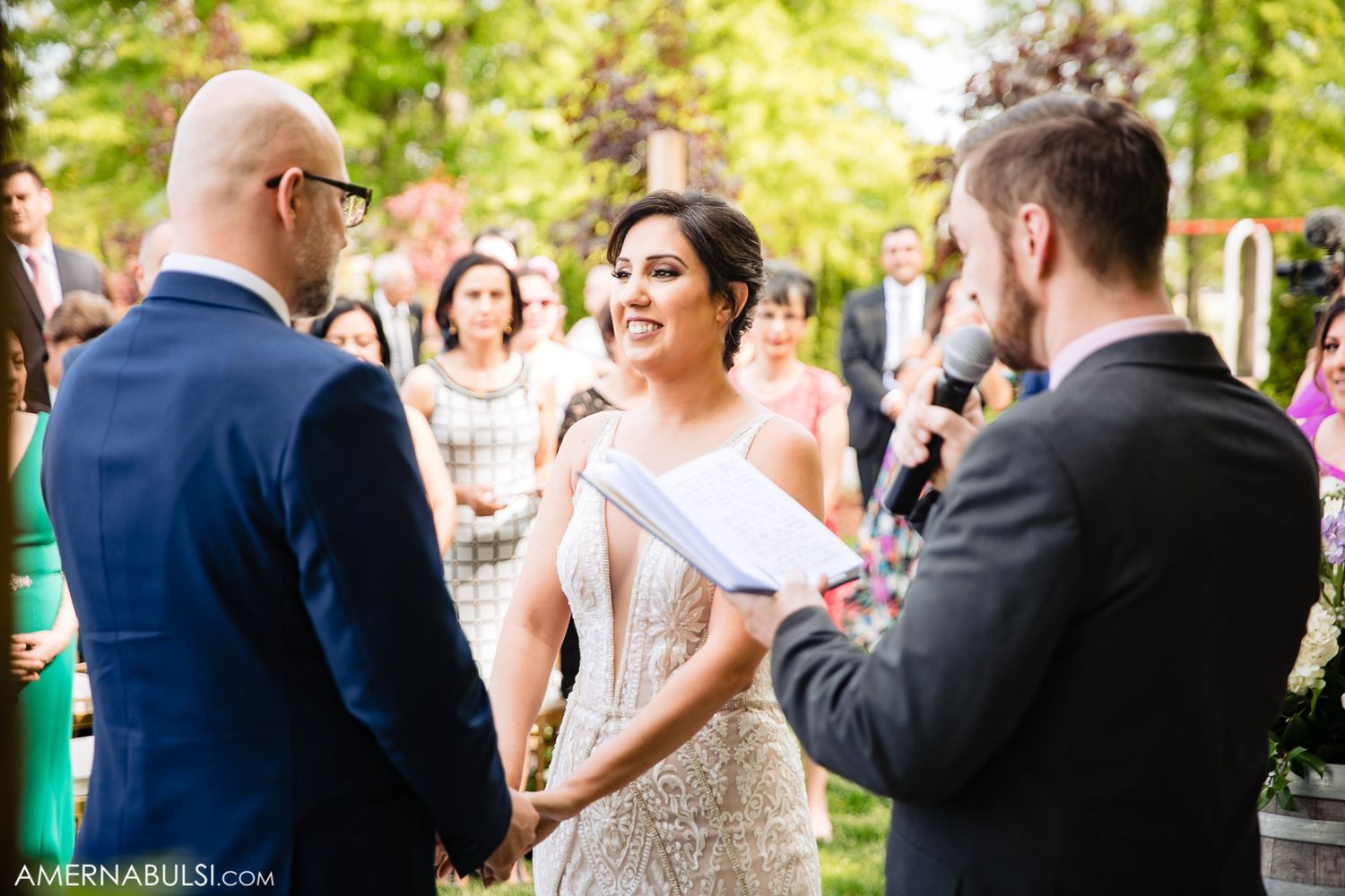 Wedding guests laugh and celebrate during an outdoor reception under green trees.