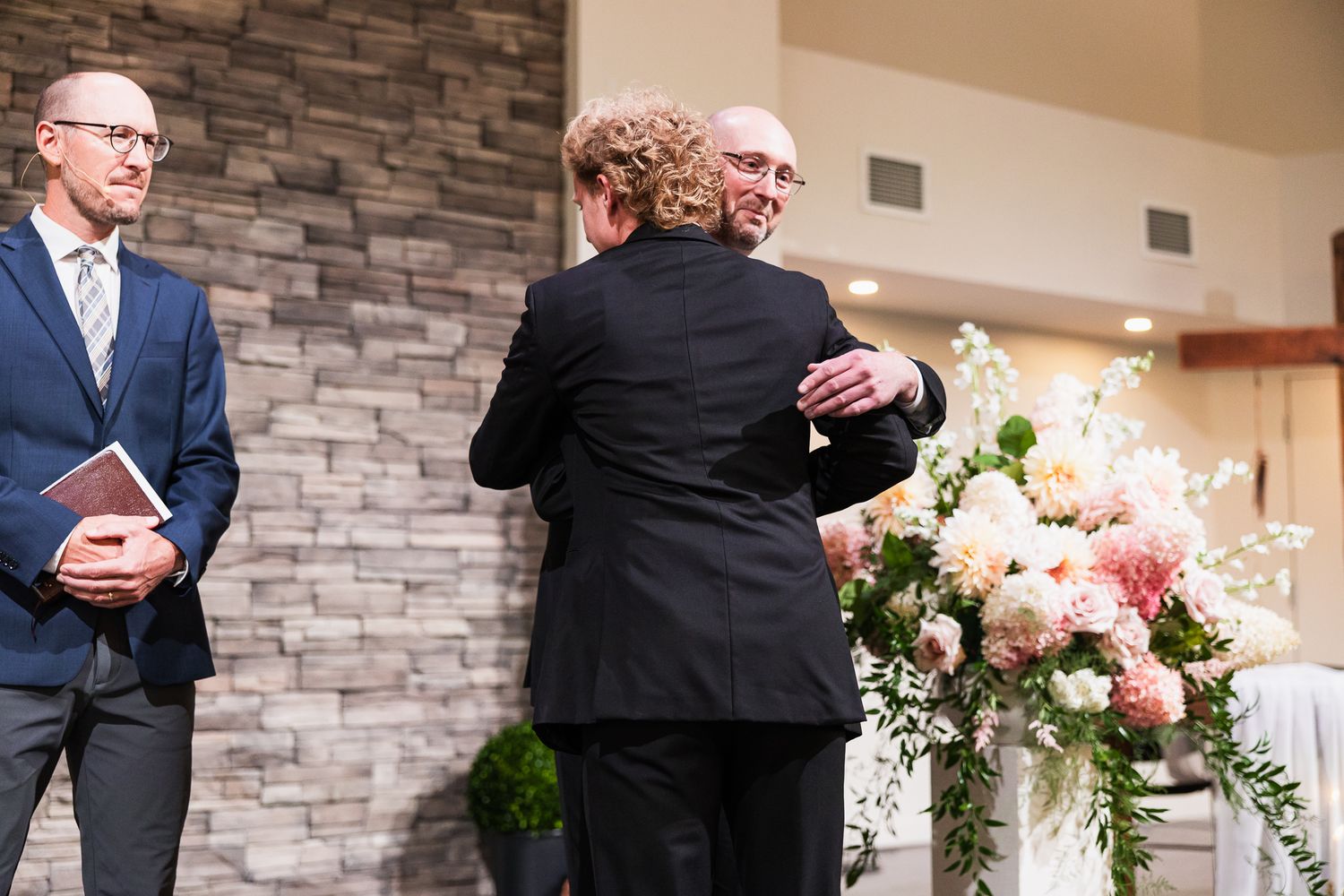 Emotional embrace during wedding ceremony with stone wall backdrop and floral arrangements.