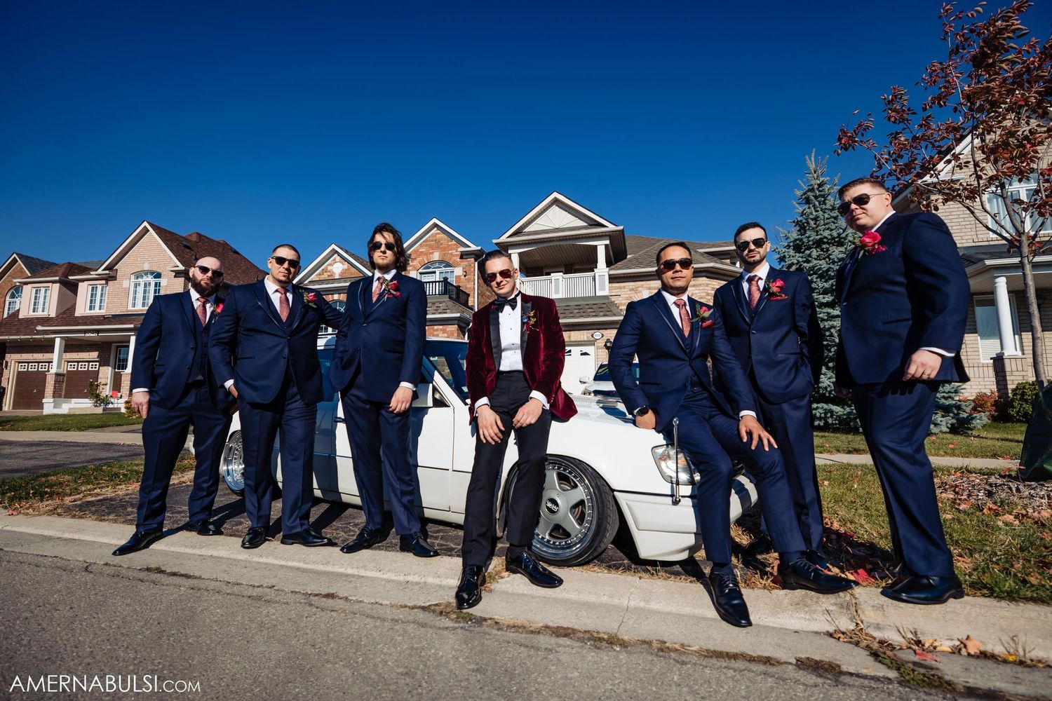 Groomsmen in navy suits pose casually around white car outside suburban homes.
